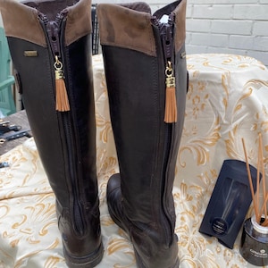 May include: A pair of tall dark brown leather boots featuring a full-length back zipper closure. Each zipper pull is adorned with a small, light tan leather tassel. The top cuff of each boot is a contrasting lighter brown leather material.