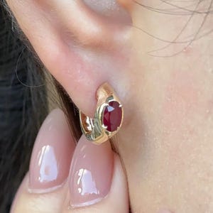 May include: A gold hoop earring featuring a single oval-cut deep red ruby gemstone set into the metal band. The earring is designed as a small, thick huggie style that sits closely against the earlobe, showcasing a polished metallic finish.