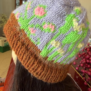 May include: A hand-knitted beanie featuring a brown ribbed brim and a light purple crown. The crown displays a colorful intarsia pattern of bright green stems and leaves with small pink and yellow flower accents throughout the knit fabric.