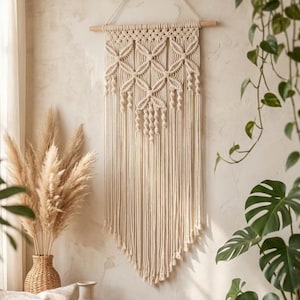May include: Cream-coloured macrame wall hanging with geometric patterns and fringe. The textile art piece is suspended from a wooden dowel, adding a touch of bohemian decor. The wall hanging is displayed near a vase of dried pampas grass.