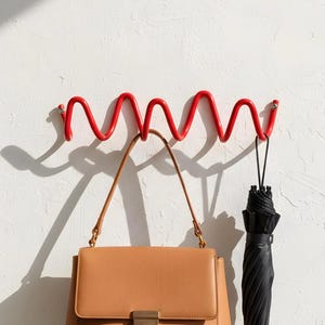 Può includere: Un attaccapanni da parete in metallo rosso con un design ondulato. Una borsa in pelle marrone e un ombrello nero sono appesi all'attaccapanni. L'attaccapanni è montato su un muro bianco.