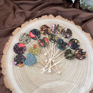 May include: A collection of tea pops on wooden skewers, arranged on a rustic wooden slice. The tea pops are round and heart-shaped, featuring various dried fruits, flowers, and herbs. The background includes a brown fabric and floral arrangements.