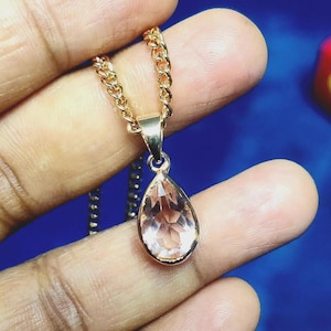 Puede incluir: Un collar dorado con un colgante en forma de lágrima. El colgante presenta una piedra preciosa rosa claro facetada. La cadena es de estilo eslabón de acera. El fondo es azul liso.