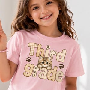 Puede incluir: Una camiseta de manga corta de color rosa claro con un estampado gráfico que incluye el texto Third grade en una fuente a cuadros beige. El diseño presenta una ilustración de un pequeño gatito atigrado marrón y huellas de patas oscuras.