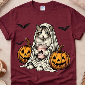 May include: A maroon short-sleeve t-shirt featuring a graphic of a cat wearing a white ghost sheet, holding a pink drink with a pumpkin icon. The cat is flanked by two orange jack-o'-lanterns and three small black bats flying in the background.