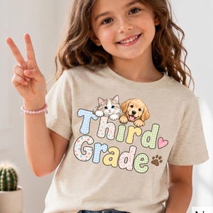 Puede incluir: Una camiseta de color natural con un gráfico de un gatito y un cachorro sobre el texto "Third Grade" en un patrón multicolor, a cuadros y floral. El diseño incluye un pequeño corazón rosa y una huella de pata marrón.