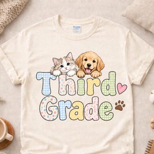 Puede incluir: Una camiseta de color natural con un gráfico de un gatito y un cachorro de golden retriever sobre el texto Third Grade. Las letras están rellenas con patrones de colores pastel, incluyendo cuadros vichy y flores, junto a un pequeño corazón rosa y una huella marrón.