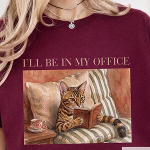 May include: Maroon-colored t-shirt featuring the text "I'LL BE IN MY OFFICE" above a watercolor illustration of a cat reading a book. The cat is brown and black, sitting on a striped pillow. A teacup sits on a small table.