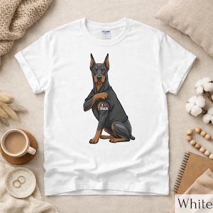 May include: White t-shirt featuring a graphic of a Doberman Pinscher with a tattoo that says "I ❤️ DAD". The dog is black and brown, with a red heart. The shirt is a Gildan brand.