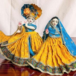 May include: Two decorative figurines of deities, one with a large headdress and the other with a blue veil. Both wear yellow skirts with ornate trim and blue tops. The figurines are set against a neutral background.
