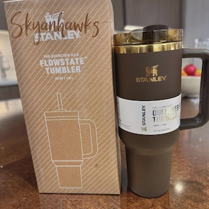 以下が含まれることがあります： 茶色のスタンレーFlowstateタンブラー。ハンドルと金色の蓋が付いています。横には段ボール箱があります。箱には「Skyanhawks」の文字とタンブラーの絵が描かれています。タンブラーは1.18Lです。