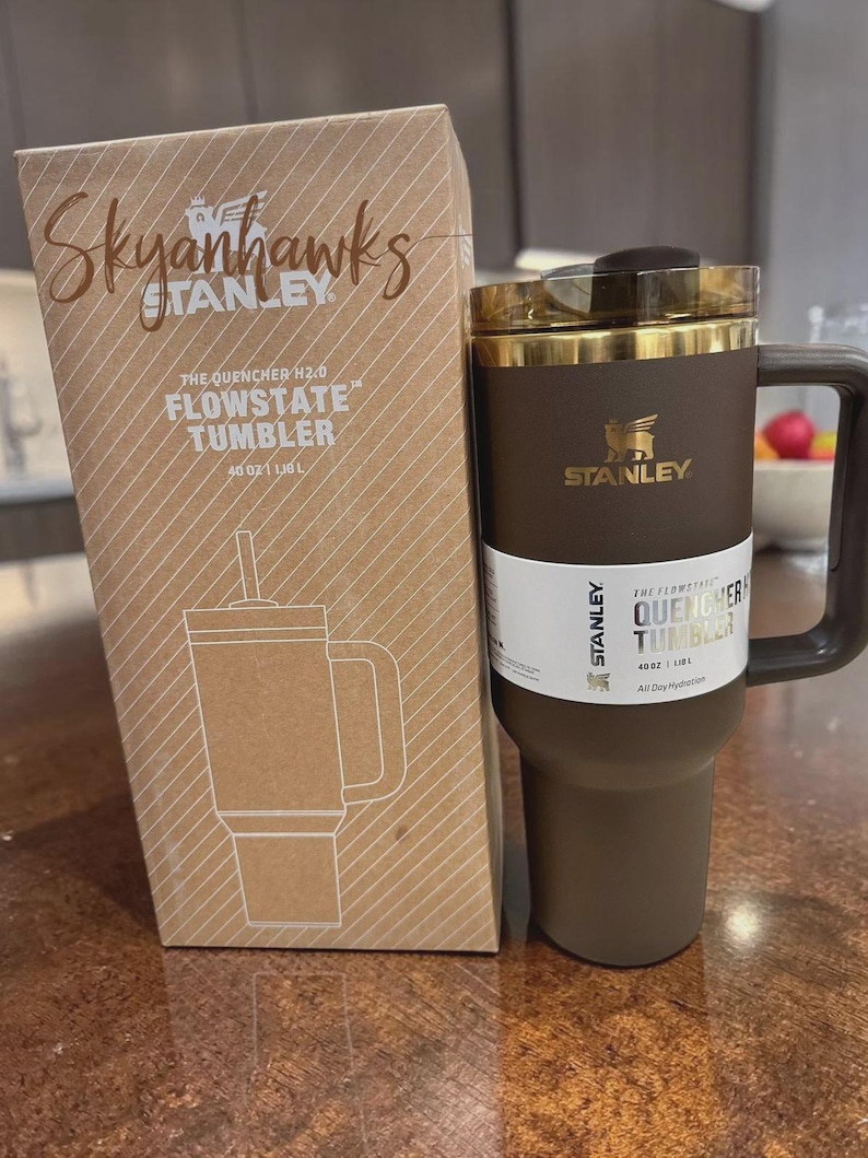 以下が含まれることがあります： 茶色のスタンレーFlowstateタンブラー。ハンドルと金色の蓋が付いています。横には段ボール箱があります。箱には「Skyanhawks」の文字とタンブラーの絵が描かれています。タンブラーは1.18Lです。