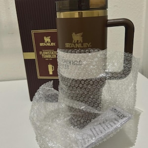以下が含まれることがあります： ハンドル、ゴールドのアクセント、濃い茶色の蓋が付いた、茶色のスタンレーフロー状態タンブラー。 タンブラーはプチプチで包まれ、スタンレーのロゴと製品情報が記載された茶色の箱の隣に置かれています。