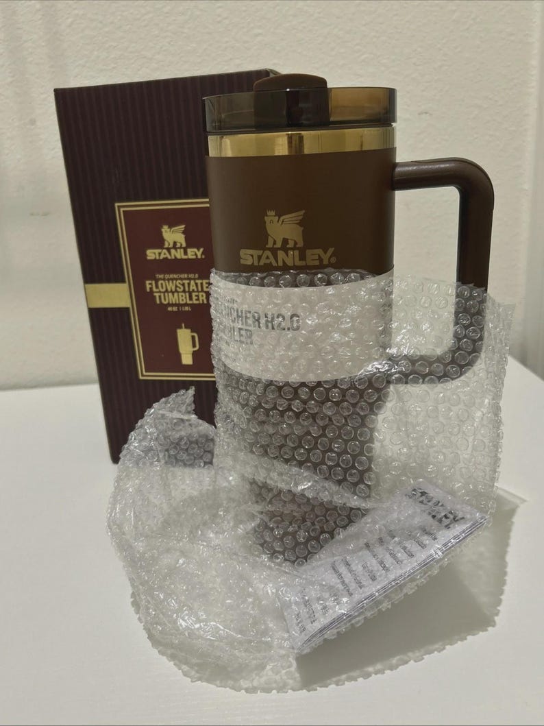 以下が含まれることがあります： ハンドル、ゴールドのアクセント、濃い茶色の蓋が付いた、茶色のスタンレーフロー状態タンブラー。 タンブラーはプチプチで包まれ、スタンレーのロゴと製品情報が記載された茶色の箱の隣に置かれています。
