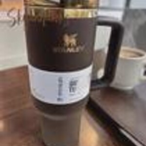 以下が含まれることがあります： ゴールドの縁と黒いハンドルが付いた、濃い茶色のスタンレーのトラベルマグ。マグにはスタンレーのロゴと白いラベルが付いています。飲み物を温かくまたは冷たく保つように設計されています。マグの高さは約18cmです。