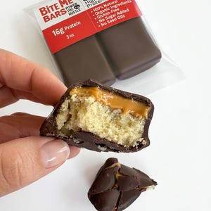 May include: A close-up of a protein bar cut in half, revealing a layered interior. The bar has a light-colored, cake-like base, a caramel layer, and a dark chocolate coating. The packaging reads "Bite Me Bars" and "Peanut Butter Caramel Protein Bites."
