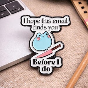 May include: A black and white enamel pin with the text "I hope this email finds you before I do." The pin features a light blue frog with a pink knife. The pin is on a beige surface next to a laptop.