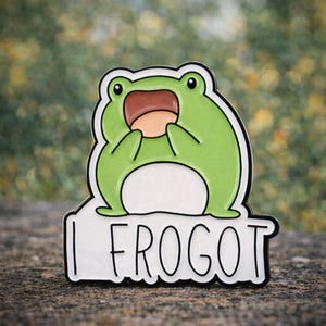 May include: A green frog enamel pin with a white border and the text "I FROGOT". The frog has a surprised expression with its mouth open. The pin is set against a blurred green and brown background.