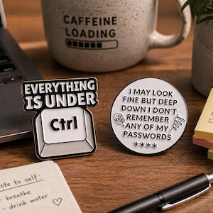May include: Two enamel pins with humorous text. One pin reads "Everything is Under Ctrl" with a keyboard key design. The other pin says "I may look fine but deep down I don't remember any of my passwords". A coffee mug and laptop are in the background.