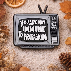 May include: A black and gray enamel pin in the shape of a vintage television set. The pin features the text "YOU ARE NOT IMMUNE TO PROPAGANDA" in white lettering. The pin is surrounded by fall-themed decorations.