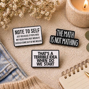 May include: Three black and white enamel pins with humorous phrases. One reads "NOTE TO SELF," another says "THE MATH IS NOT MATHING," and the third states "THAT'S A TERRIBLE IDEA WHEN DO WE START." The pins are arranged on a neutral surface with a lit candle and a notebook.