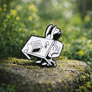 Puede incluir: Un pin de esmalte en blanco y negro que representa a una persona en un escritorio con un portátil y un ratón. La persona está desplomada sobre el escritorio, con las manos en la cabeza. El pin está sobre un fondo verde borroso.