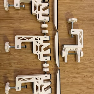May include: A collection of white 3D-printed clamps and components, including adjustable screw clamps and black-edged panels. The clamps have a geometric design and are arranged on a wooden surface.