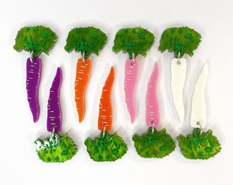 May include: Nine pairs of carrot earrings with green tops. The carrots are made of acrylic and come in a variety of colors, including purple, orange, pink, and white.