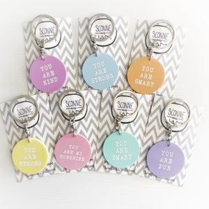 May include: A collection of colorful keychains with silver rings. Each round keychain has a different pastel color and a positive affirmation in white text, such as "You Are Kind" and "You Are Strong". The keychains are displayed on chevron patterned cards.