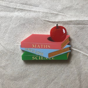 Peut inclure: Une pile de trois livres avec les titres "MATHS", "ENGLISH" et "SCIENCE" en lettres dorées. Une pomme rouge est posée sur les livres. Les livres sont roses, bleu clair et verts. Le fond est un tissu beige clair.