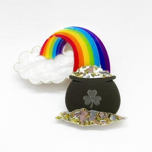 May include: A colorful brooch featuring a rainbow arching over a white cloud and a black pot filled with gold. The pot has a silver shamrock design. The rainbow has red, orange, yellow, green, blue, and purple stripes.
