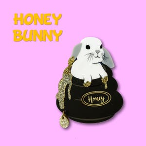 May include: A white rabbit with grey ears sits in a black pot of honey, with gold glitter accents. The word "Honey" is on the pot. The text "HONEY BUNNY" is in yellow letters on a pink background.