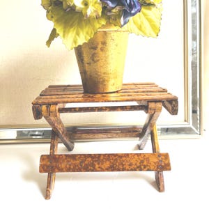 May include: A small, folding wooden stool with a mottled brown finish, featuring a slatted top and cross-legged base. Resting on the stool is a gold-colored pot containing a bouquet of green and dark blue artificial flowers.