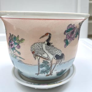 May include: A ceramic planter with a matching saucer featuring a pale pink background, hand-painted white cranes with black and red accents, and purple floral motifs. The bowl-shaped vessel has a wide rim and a smooth, glossy finish throughout.