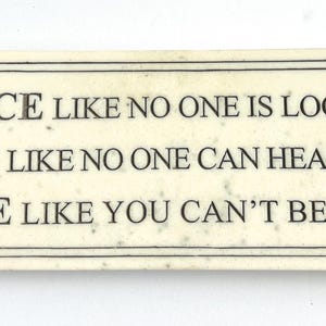 May include: A rectangular off-white plaque with a speckled texture and a thin black border. The text reads: DANCE LIKE NO ONE IS LOOKING, SING LIKE NO ONE CAN HEAR YOU, LOVE LIKE YOU CAN'T BE HURT. The lettering is in a dark, serif font.