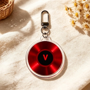 May include: A red and black acrylic keychain with a silver clasp. The keychain has a circular design with a red background and a black center featuring the letter "V".