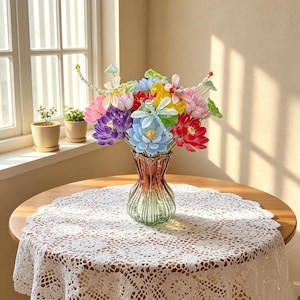 Könnte beinhalten: Ein farbenfroher Strauß künstlicher Blumen in einer dekorativen Glasvase. Das Arrangement umfasst rosa, lila, blaue, gelbe und rote Blüten. Die Vase steht auf einem runden Holztisch, der mit einer weißen Spitzendecke bedeckt ist.