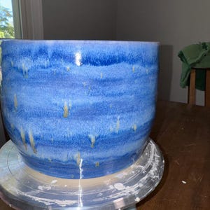 Puede incluir: Una maceta cilíndrica de cerámica con un esmalte brillante que presenta bandas horizontales de azul cobalto profundo, azul cielo más claro y sutiles vetas de goteo en color crema. El recipiente tiene un borde liso y redondeado.