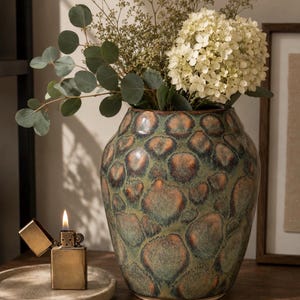 May include: A ceramic vase with a rounded body and a mottled glaze featuring shades of green, brown, and copper. The vase contains a bouquet of white hydrangea flowers and green eucalyptus branches, with a brass lighter nearby.