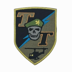 May include: Embroidered patch with a shield shape, featuring a skull wearing a green beret with a shamrock. The patch includes the letters "T" and a lightning bolt, and a helicopter silhouette. The patch has a black, olive green, and yellow color scheme.