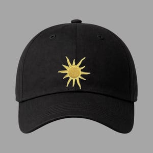 May include: Black baseball cap with a gold embroidered sun design. The sun has a textured center and radiating lines. The cap has a curved brim and a button on top.