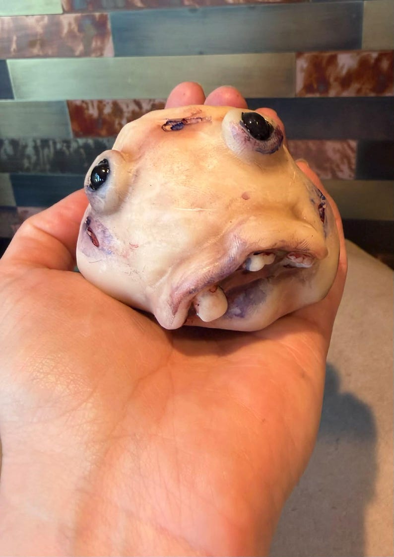 K&ouml;nnte beinhalten: Eine blasse, fleischfarbene, abgerundete Skulptur mit strukturierter Oberfl&auml;che, zwei hervorstehenden schwarzen, perlenartigen Augen, einem kleinen Mund mit sichtbaren wei&szlig;en Z&auml;hnen und dezenter violetter und roter Schattierung auf der Form.