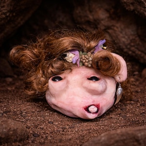 Puede incluir: Una cabeza de muñeca esculpida con piel pálida, ojos oscuros y un solo diente visible en la boca. Presenta cabello castaño ondulado adornado con pequeñas flores moradas y blancas y bayas marrones. Un pendiente plateado en forma de ala cuelga de una oreja.