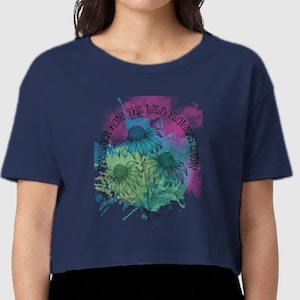 Puede incluir: Una camiseta corta de color azul marino con mangas cortas y un estampado gráfico de flores equináceas azules y verdes. El diseño incluye un fondo estilo acuarela en rosa y morado y el texto "Wonder how the wild flowers grow".