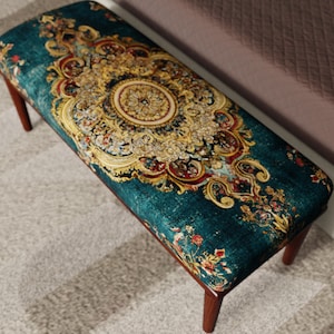 Puede incluir: Banco rectangular con tapizado turquesa con un diseño floral ornamentado en dorado, rojo y crema. El banco tiene cuatro patas de madera oscura y está sobre una alfombra clara. Mide aproximadamente 120 cm de largo.