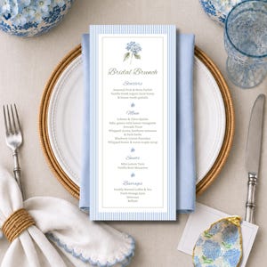 May include: A rectangular menu card with a light blue and white vertical striped border and a small blue hydrangea illustration at the top. The text reads Bridal Brunch, Starters, Main, Sweets, and Beverages, listing various food and drink items.