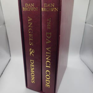 Könnte beinhalten: Ein Schuber mit zwei Büchern von Dan Brown. Die Bücher sind in tiefem Burgunderrot gebunden und mit goldfarbenen Buchstaben versehen. Die Titel lauten "Angels & Demons" und "The Da Vinci Code".
