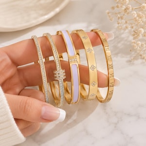 Könnte beinhalten: Ein Set aus fünf goldfarbenen Metallarmreifen. Der erste ist mit kleinen klaren Steinen besetzt, der zweite hat einen dreifachen Ringakzent mit klaren Steinen, der dritte eine hellviolette Emaille-Einlage, der vierte florale Motive und der fünfte römische Ziffern.