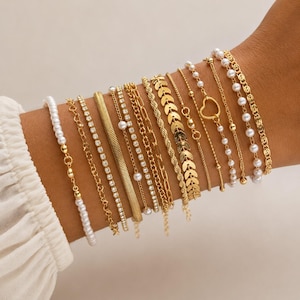 Könnte beinhalten: Eine Sammlung goldfarbener Armbänder, darunter Perlen-, Ketten- und Charm-Designs, wird an einem Handgelenk präsentiert. Die Armbänder variieren im Stil und verfügen über zarte Ketten, funkelnde Strasssteine und herzförmige Anhänger. Die Gesamtästhetik ist elegant und modisch.