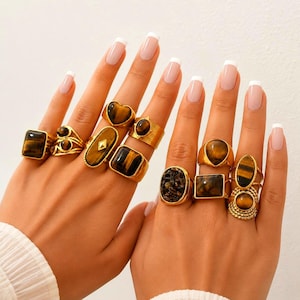 Op de afbeelding: Een collectie goudkleurige ringen met tijgeroog-edelstenen in diverse vormen, waaronder rechthoekige, ovale, hartvormige en ronde zettingen. De ringen worden aan de vingers gedragen en tonen verschillende ringstijlen, van gladde tot textuurafwerkingen.